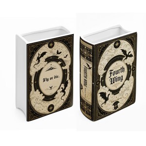 May include: A white ceramic book-shaped vase featuring black and cream illustrations of dragons, clouds, and stars. One side displays the text 'Fly or die.' while the other side features the title 'Fourth Wing' by Rebecca Yarros in black lettering.