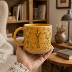 May include: Mustard yellow ceramic mug with a black sword and floral design. The mug has the text "you do not fear, you do not falter, you do not yield". The mug has a beige base and a curved handle.