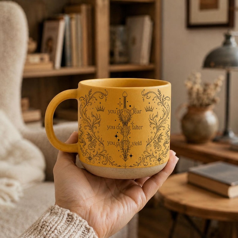 May include: Mustard yellow ceramic mug with a black sword and floral design. The mug has the text "you do not fear, you do not falter, you do not yield". The mug has a beige base and a curved handle.