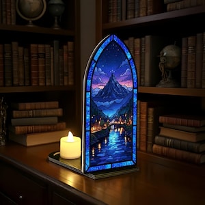 Pu&ograve; includere: Un pannello decorativo in vetro colorato raffigurante un paesaggio montano con un fiume e una citt&agrave;, illuminato da una calda candela a LED. Il pannello ha la forma di un arco gotico, con un bordo blu e una base nera. La scena include un cielo stellato.