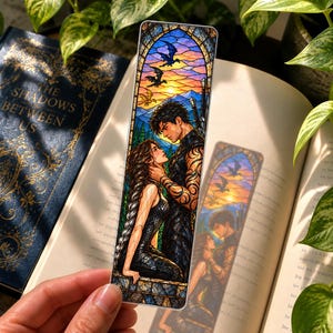 May include: A rectangular bookmark featuring a stained glass style illustration of two figures facing each other. The background shows a sunset sky with flying dragons above mountains. The figures are depicted with intricate line work and vibrant colours.