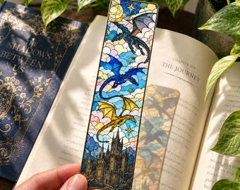 May include: A rectangular bookmark featuring a stained glass style illustration of three dragons in flight above a dark castle. The design includes a black dragon, a blue dragon, and a yellow dragon set against a sky of blue, pink, and white clouds.