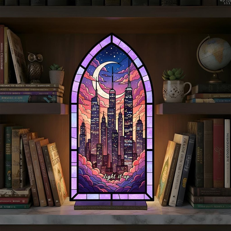 May include: A stained glass window-shaped lamp featuring a cityscape silhouette under a crescent moon and starry sky. The artwork is framed in purple and the text "light it up" is at the bottom. The lamp is set on a wooden base.