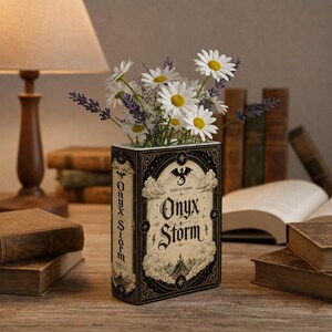Pu&ograve; includere: Un vaso rettangolare a forma di libro con un design di copertina color crema e nero con il titolo Onyx Storm di Rebecca Yarros, decorato con illustrazioni di nuvole, un drago e stelle, riempito con margherite bianche e rametti di lavanda viola.