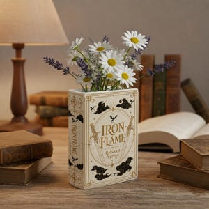Pu&ograve; includere: Un vaso in ceramica rettangolare a forma di libro con il titolo Iron Flame di Rebecca Yarros stampato sul davanti. Il vaso &egrave; color crema con accenti decorativi dorati e neri, tra cui spade, nuvole e un drago, riempito con margherite bianche e lavanda viola.