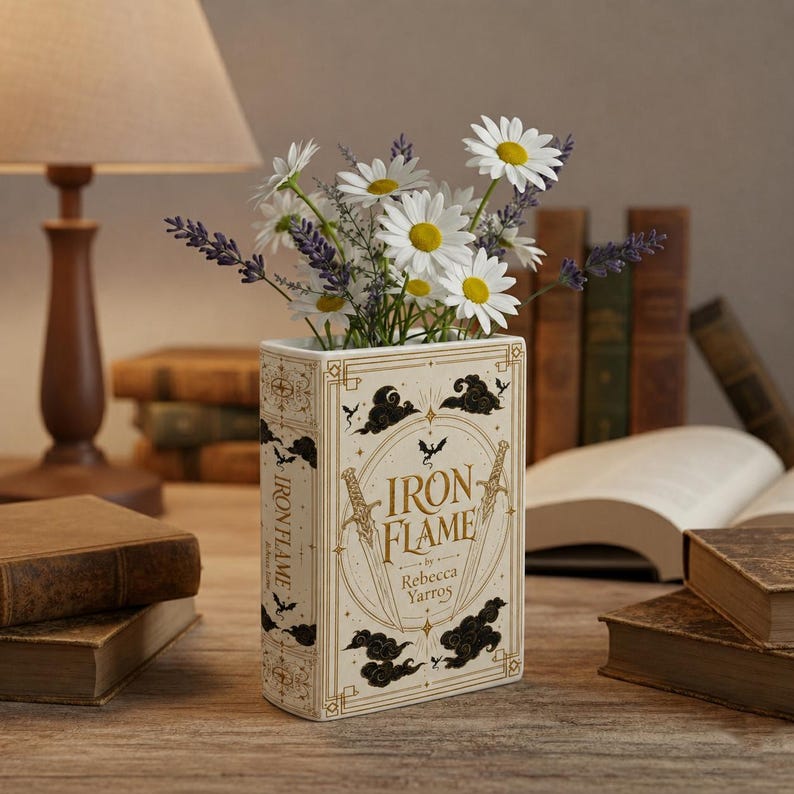 Pu&ograve; includere: Un vaso in ceramica rettangolare a forma di libro con il titolo Iron Flame di Rebecca Yarros stampato sul davanti. Il vaso &egrave; color crema con accenti decorativi dorati e neri, tra cui spade, nuvole e un drago, riempito con margherite bianche e lavanda viola.