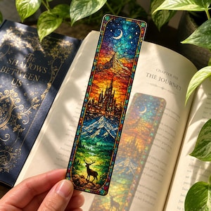 Pode incluir: Um marcador de página retangular com um design de vitral colorido. A ilustração mostra uma lua crescente e estrelas sobre uma cordilheira, um castelo alto, um pico nevado, um vale verde e a silhueta de um cervo na parte inferior.