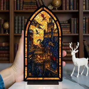May include: A stained glass panel showing a fantasy castle scene with dragons and a fiery sky. The artwork is framed in a dark border and set on a wooden base. The panel is held in a hand.