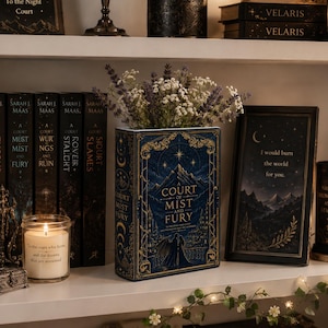 May include: A rectangular book-shaped vase in deep blue with gold foil illustrations of mountains, a castle, and two figures. The vase holds a bouquet of dried lavender and white flowers, positioned on a shelf next to books and a framed quote.