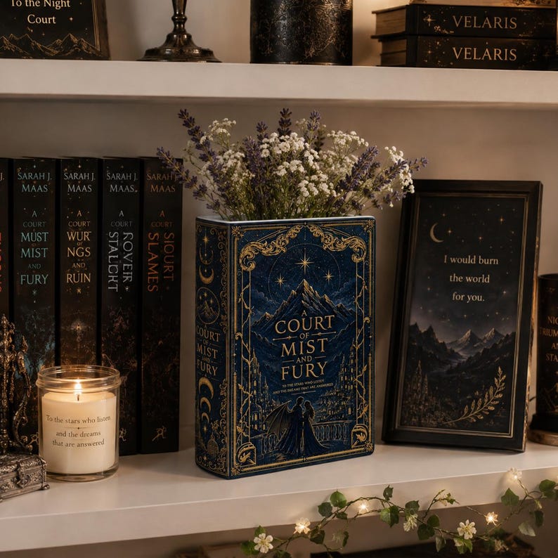 May include: A rectangular book-shaped vase in deep blue with gold foil illustrations of mountains, a castle, and two figures. The vase holds a bouquet of dried lavender and white flowers, positioned on a shelf next to books and a framed quote.