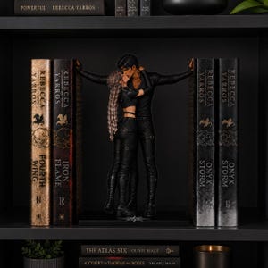 May include: A pair of black bookends featuring a couple in a romantic embrace, positioned between two sets of books. The figures are dressed in black outfits, with the person on the left having long braided hair. The books include titles by Rebecca Yarros.