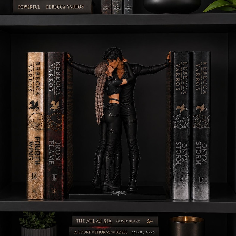 May include: A pair of black bookends featuring a couple in a romantic embrace, positioned between two sets of books. The figures are dressed in black outfits, with the person on the left having long braided hair. The books include titles by Rebecca Yarros.