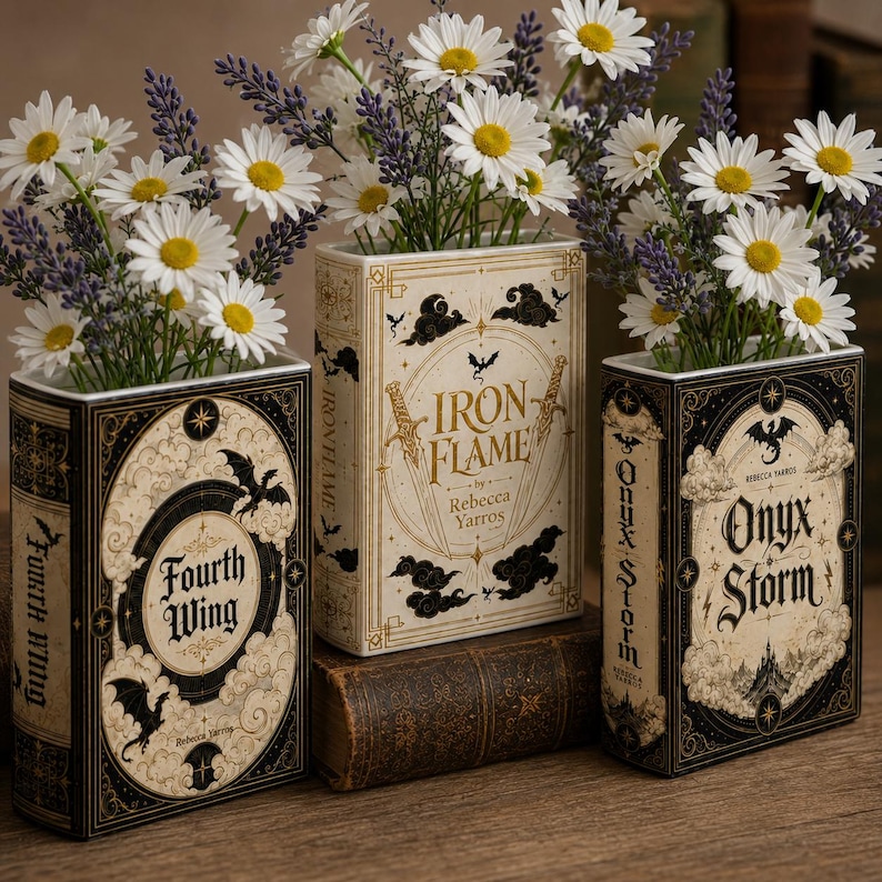 Pu&ograve; includere: Tre vasi rettangolari a forma di libro con disegni neri e crema e dettagli dorati. Il vaso a sinistra riporta la scritta Fourth Wing, quello centrale Iron Flame e quello a destra Onyx Storm. Ogni vaso &egrave; riempito con margherite bianche e lavanda viola.