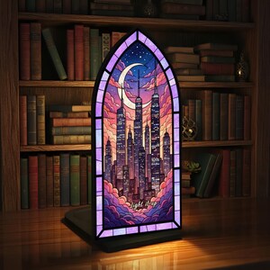 May include: A stained glass window-shaped lamp featuring a cityscape silhouette under a crescent moon and starry sky. The lamp has a purple border and the words "light it up". The lamp is on a black base, with a bookshelf in the background.