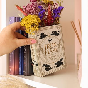 May include: A hollowed-out book titled Iron Flame by Rebecca Yarros, functioning as a planter filled with dried yellow, purple, and red flowers. The book cover features gold and black line art of swords, clouds, and a dragon silhouette.