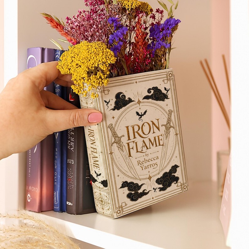 May include: A hollowed-out book titled Iron Flame by Rebecca Yarros, functioning as a planter filled with dried yellow, purple, and red flowers. The book cover features gold and black line art of swords, clouds, and a dragon silhouette.