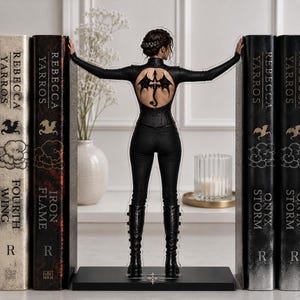 May include: A clear acrylic standee featuring a figure in a black long-sleeved top with a circular back cutout displaying a dragon tattoo, black pants, and black boots, positioned between two large book-shaped decorative bookends.
