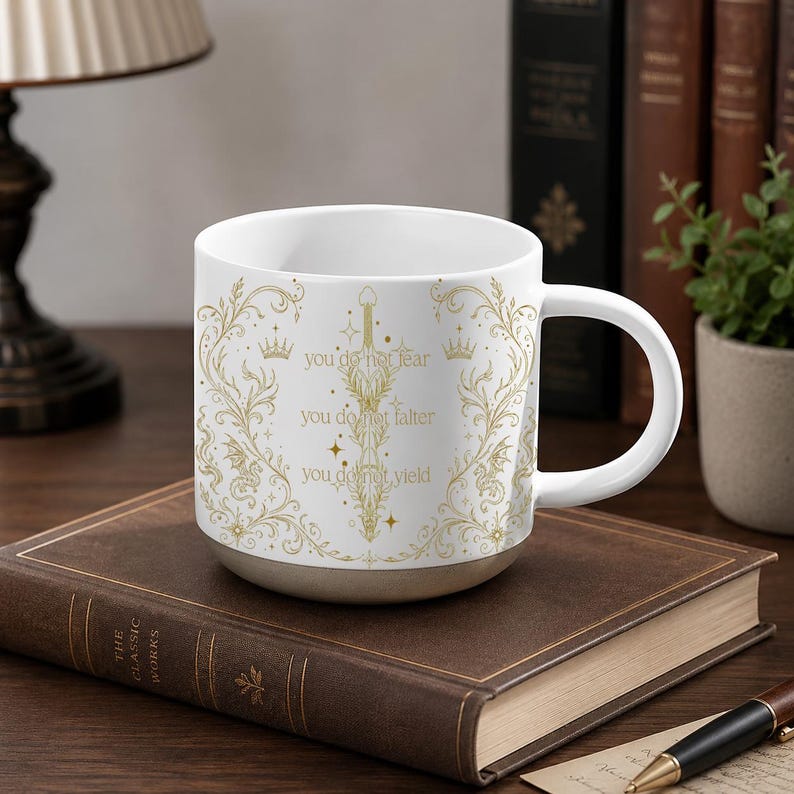 May include: White ceramic mug with a gold floral design and the text "you do not fear, you do not falter, you do not yield." The mug has a beige base and a white handle. It sits on a brown book.