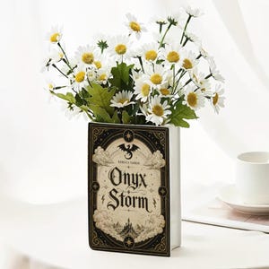 Puede incluir: Un florero rectangular de cer&aacute;mica blanca con forma de libro, que presenta un dise&ntilde;o de portada en negro y crema con el texto Onyx Storm y Rebecca Yarros, lleno de un ramo de margaritas blancas con centros amarillos y hojas verdes.