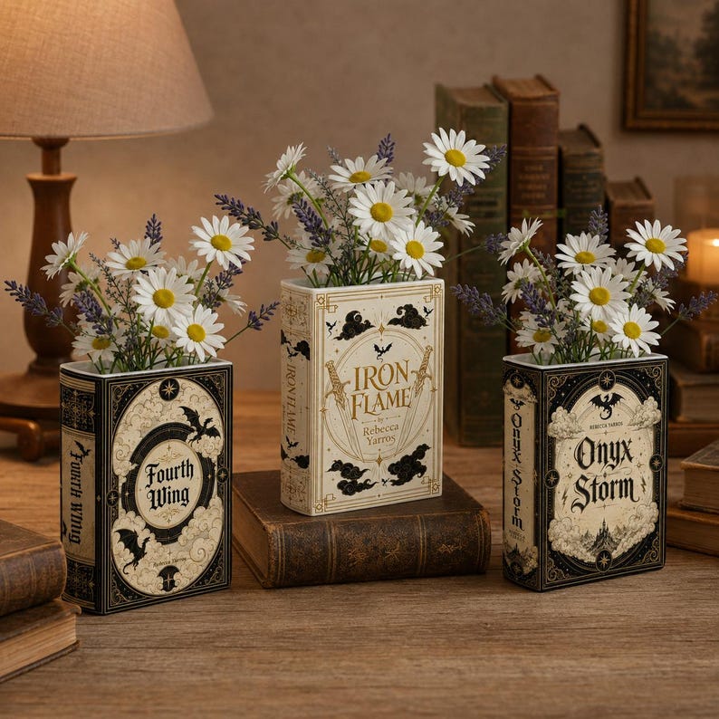 Pu&ograve; includere: Tre vasi rettangolari a forma di libro con copertine decorative intitolate Fourth Wing, Iron Flame e Onyx Storm. Ogni vaso &egrave; riempito con fiori bianchi simili a margherite e rametti di lavanda viola, con intricate illustrazioni grafiche nere e oro.