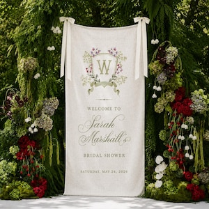 May include: A rectangular off-white fabric banner featuring a floral crest with the letter W, followed by the text Welcome to Sarah Marshall's Bridal Shower and the date Saturday, May 24, 2026. The banner is suspended by two cream-colored ribbons.