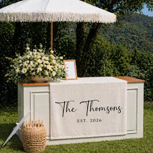 May include: A cream-colored outdoor bar with a white fringed umbrella, floral arrangement, and a sign that reads "The Thomsons EST. 2026." A small basket and parasol are on the grass in front of the bar. The background features a lush green hillside.