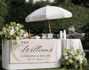 Könnte beinhalten: Eine weiße, ovale, geriffelte Bartheke, die mit einem beigen Stoffbanner mit dem Text THE Williams CATHERINE & MILLER 07.31.2026 bedeckt ist. Ein weißer Sonnenschirm mit Fransen steht dahinter, ergänzt durch Blumenarrangements und einen Getränkespender.