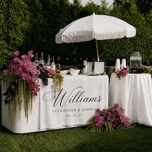 May include: A white rectangular bar table with a custom fabric cover featuring the text THE Williams ALEXANDER & SOPHIA 06.21.27, adorned with purple orchids and dark calla lilies, accompanied by a white fringed umbrella and a matching round table.