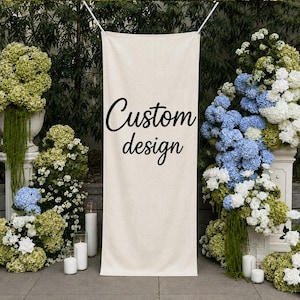 May include: A vertical rectangular beige fabric banner suspended by white strings, featuring the black text Custom design printed in a cursive script font, positioned centrally between two large floral arrangements of blue and green hydrangeas.