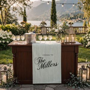 May include: A wooden bar with a striped runner that reads "Cheers to The Millers." The bar is set with champagne, wine glasses, and a floral arrangement. Lanterns and greenery flank the bar, with a scenic background.