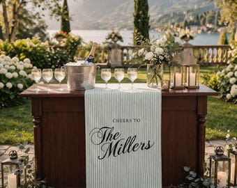 May include: A wooden bar with a striped runner that reads "Cheers to The Millers." The bar is set with champagne, wine glasses, and a floral arrangement. Lanterns and greenery flank the bar, with a scenic background.