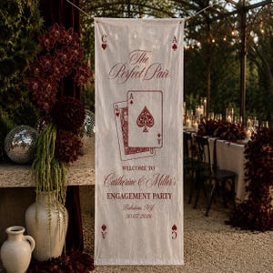 May include: A vertical off-white fabric banner featuring red printed text and graphics. The design includes the phrase The Perfect Pair, an illustration of a Queen and Ace of Spades playing card, and text reading Welcome to Catherine & Miller's Engagement Party, Baladwin, NJ, 30.07.2026, with small spade and heart symbols in the corners.