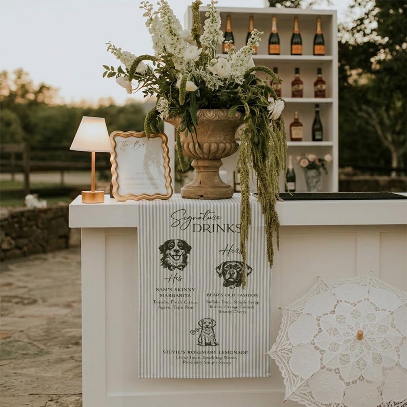 Peut inclure: Un bar blanc avec un arrangement floral, un panneau dor&eacute; et une enseigne "Signature Drinks". Le panneau pr&eacute;sente des illustrations de chiens et des noms de boissons. Un parasol en dentelle blanche est &agrave; droite.