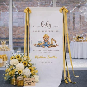 May include: A white banner with the text "baby UNDER CONSTRUCTION" and "WELCOME TO THE BABY SHOWER IN HONOR OF Mathew Smiths APRIL 17, 2026". The banner features a teddy bear in overalls and construction vehicles. Yellow ribbons and a floral arrangement are also present.