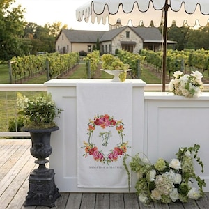 May include: A white linen towel with a floral wreath design and the initials "J" is displayed on a white counter. The towel is decorated with pink, red, and orange flowers. The words "SAMANTHA & MATHEO" are printed below the floral design.