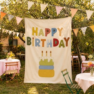 May include: A large off-white fabric banner featuring the text HAPPY BIRTHDAY in colorful, patterned fabric letters. Below the text is a three-tiered cake illustration with three lit candles, all rendered in various fabric textures and colors.