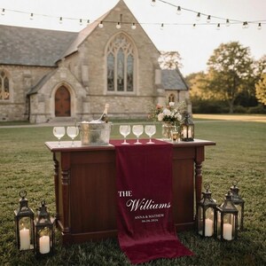 May include: A dark wood bar with a burgundy runner, wine glasses, and a champagne bottle in an ice bucket. The runner has the text "The Williams" and a date. Decorative lanterns with lit candles are in the foreground.
