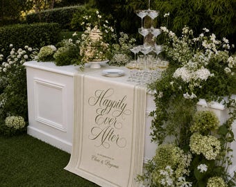 May include: A white table runner featuring the text "Happily Ever After" and "In celebration of Olivia & Benjamin" in elegant dark script, accented by vertical lines, displayed on a white table with a champagne tower and white floral arrangements.