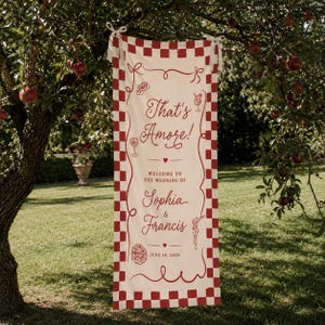 May include: A rectangular fabric banner with a red and white checkered border. The center features red text reading That's Amore! Welcome to the wedding of Sophia & Francis, June 10, 2025, accompanied by red line art illustrations of wine, pasta, and pizza.
