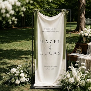 May include: A vertical off-white fabric wedding welcome sign suspended from a black metal frame, featuring the text "WELCOME TO THE WEDDING OF HAZEL & LUCAS" with the date "September 27, 2026" and "Denver, CO," accented by two olive green satin ribbons.
