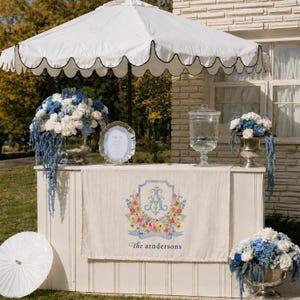 May include: A white outdoor bar with a scalloped umbrella, floral arrangements, and a banner with a floral crest and the text "the arndersons". The bar has a water dispenser and a decorative sign.