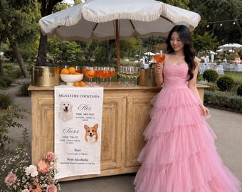 May include: A woman in a pink, layered gown stands near a bar with a white umbrella. The bar has a sign that reads "Signature Cocktails." Oranges and cocktails are on the bar. The sign lists "His" and "Hers" cocktails. The woman holds a cocktail.