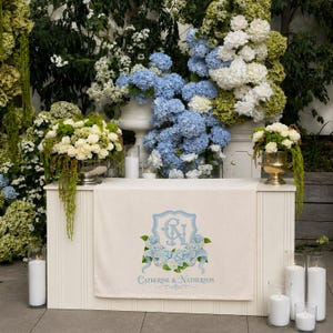 May include: A white table with a cream-colored runner featuring a light blue crest with the initials "C & N" and the names "Catherine & Natherson". The table is decorated with white and blue hydrangeas and candles, creating a wedding or event theme.