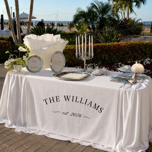 May include: A white table runner featuring the black printed text THE WILLIAMS and est 2026. The table is decorated with a silver candelabra, two silver oval frames, a silver tray with a card, a white floral arrangement, and a white candle.