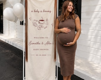 Peut inclure: Une grande bannière rectangulaire en tissu beige suspendue à un cadre métallique blanc avec des rubans marron. La bannière présente un texte marron foncé indiquant a baby is brewing, WELCOME TO Samatha & Alex's BABY SHOWER et 05.16.2026.