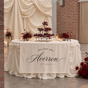May include: A round table covered with a cream-colored cloth featuring a runner printed with the text MR AND MRS. Averron EST. 2026. The table is decorated with a tiered silver stand filled with red grapes and two floral arrangements of red roses.