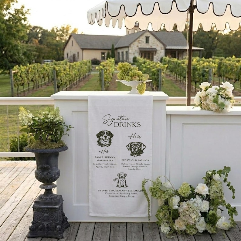 Peut inclure: Un bar blanc avec un parasol &agrave; rayures noires et blanches, un vignoble en arri&egrave;re-plan et une serviette en lin blanche avec les mots "Signature Drinks" et des illustrations de chiens. La serviette liste des recettes de boissons.