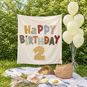 May include: A cream-colored fabric banner featuring the text Happy Birthday and the number 2 in various patterned, multi-colored fabric letters and numbers. The banner is displayed outdoors with a bunch of pale yellow balloons and a woven basket.