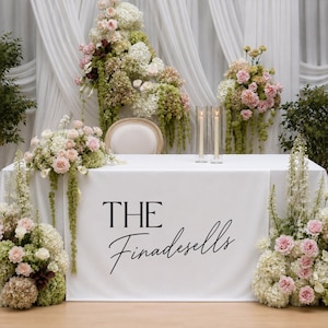 May include: A white rectangular table covered with a white cloth featuring the black text THE Finadesells. The table is decorated with large floral arrangements of pink and white flowers and green foliage, plus two tall clear glass candle holders.