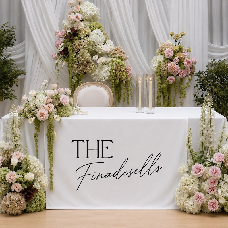 May include: A white rectangular table covered with a white cloth featuring the black text THE Finadesells. The table is decorated with large floral arrangements of pink and white flowers and green foliage, plus two tall clear glass candle holders.